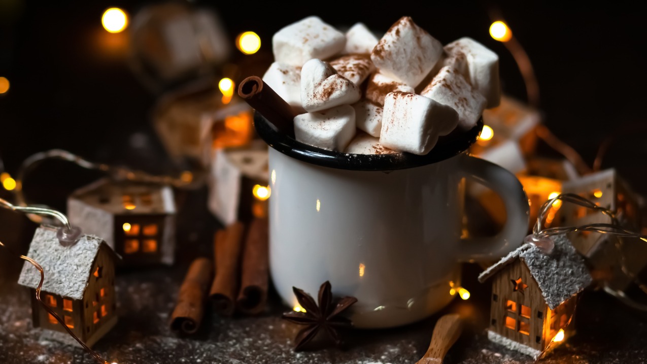 Winter hot drink: hot chocolate with marshmallows, cinnamon and anise. Christmas lights in the shape of houses as decor. Decorative snow, dark wooden background. Cozy home atmosphere. 