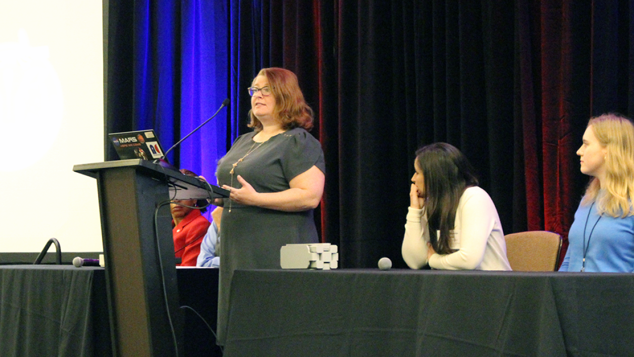 Hoover is standing in front of a podium holding a laptop computer and is speaking into a microphone while four women sitting beside her look at her.