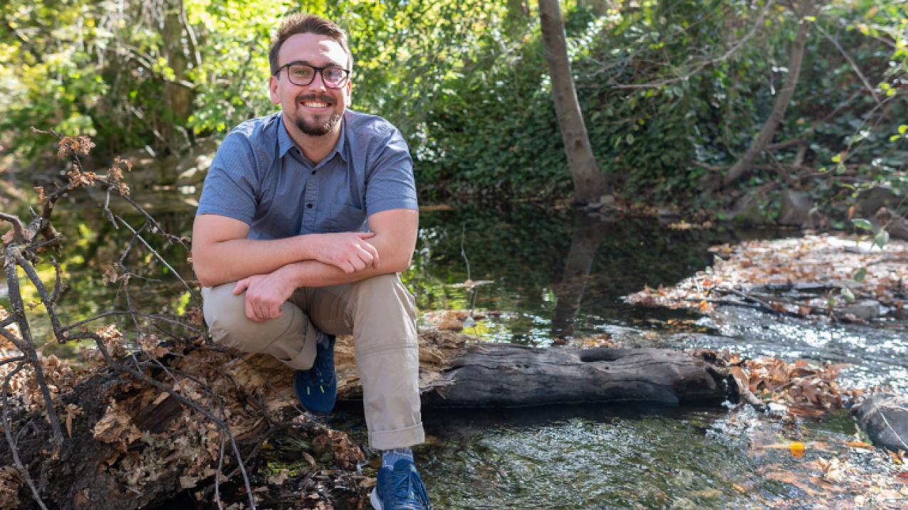 Cole wears business casual clothing and sits on a log in a wooded streamside landscape.