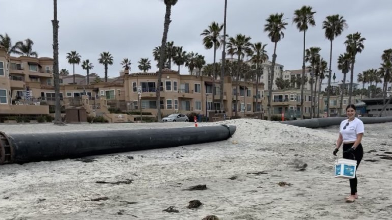 Daniela Loera stands holding a bucket next to a giant pipe running along the beach. Residential homes and palm trees are pictured in the background. 