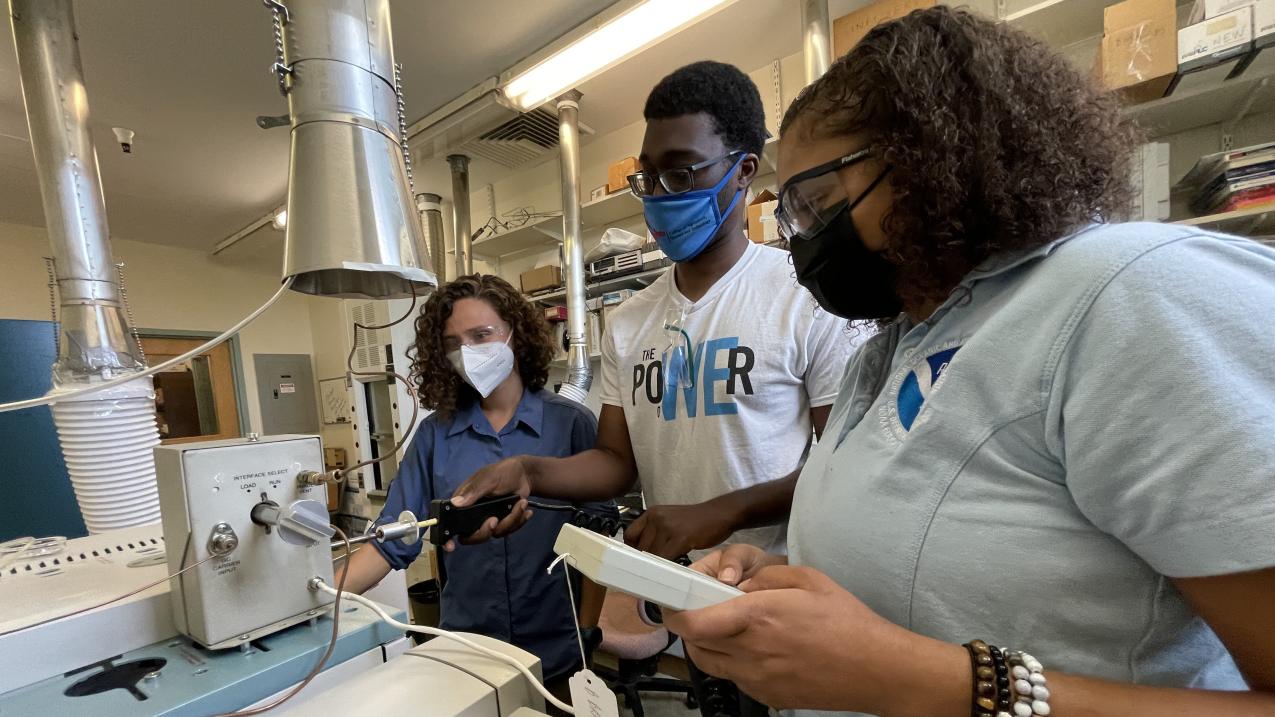 Sophia, Marcus, and Imani stand around a large piece of equipment in a lab. All three are focused on an instrument that Marcus appears to be inserting into the equipment. Imani holds another different instrument, which looks like it may have buttons. All three wear lab safety glasses and a mask of the kind that is used to prevent the spread of COVID-19. 