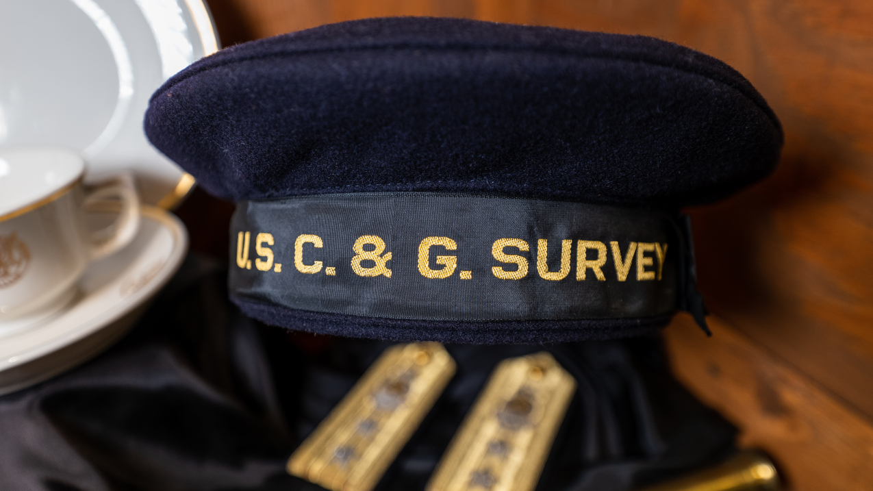 A U.S. Coast & Geodetic Survey (USC&GS) cap sits on a dark wooden background. The  black cap is shaped like a beret with a wide band at the bottom. On the band, "U.S. C. & G. SURVEY" is written in gold.