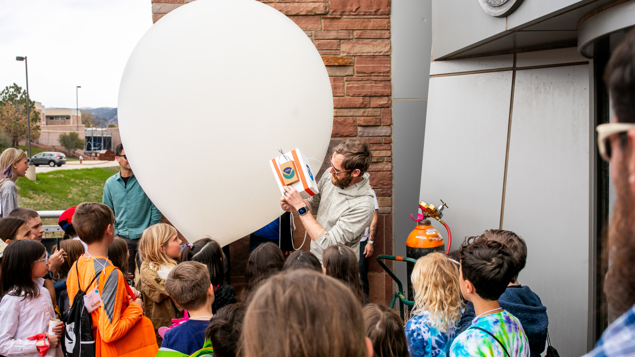 Standing in front of a group of more than a dozen children and several adults, Patrick Cullis holds up a small box with two NOAA logos that appears to contain scientific equipment. He is standing next to an inflated weather balloon and a canister of gas. The onlookers whose faces are visible appear to be watching with rapt attention.