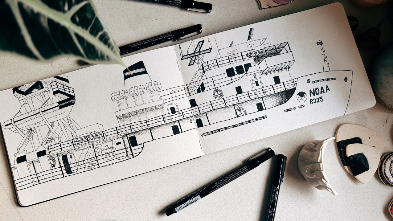 View of a pen-and-ink drawing of NOAA Ship Oscar Elton Sette across the spread of a moleskine notebook, placed on a desk and artfully surrounded by pens, ceramic decorations, and the leaf of a houseplant.