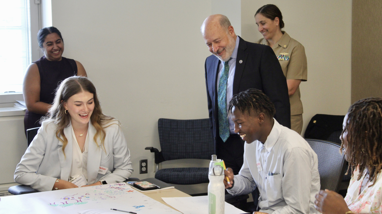 NOAA Administrator Dr. Rick Spinrad smiles as he looks over the shoulder of a teen sitting at a table. The teen, along with two other teens sitting at the table, are showing Dr. Spinrad some sketches and notes. All are smiling, as are two adults looking on from the background.