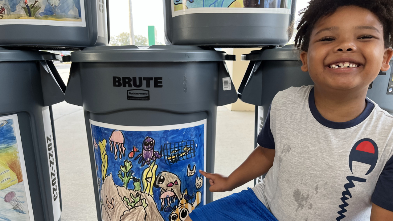 A young boy kneels next to and points to a garbage can with a large sticker of his colorful artwork on the side. The illustration appears to have been done with markers and crayons and features a variety of sea creatures with large, cute eyes as well as algae, plastic bags, and a net with fish in it. Other garbage cans featuring children’s marine debris-related art are arranged next to and stacked on top of the can he is pointing to.