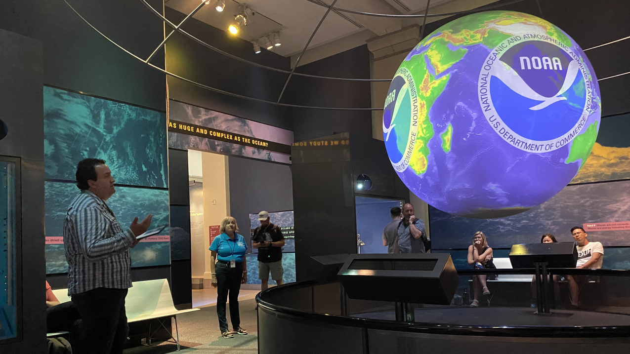 Juan Pablo Hurtado stands in a dark room in a museum with a large bright sphere hanging from the ceiling. The sphere is showing a large NOAA logo superimposed over a dataset of the Earth’s elevation and bathymetry. Several museum visitors and staff sit and stand in the background, looking at the sphere.