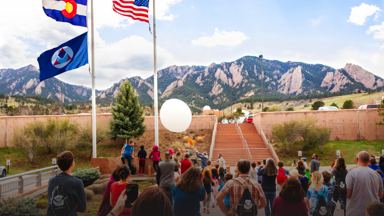 Children and adults gather to assist two scientists launching a weather balloon in windy conditions. The U.S., Colorado, and NOAA flags flutter in the breeze, framed by the Flatiron Mountains in the distance. 