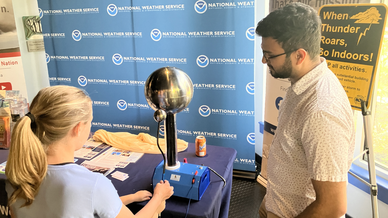 An older student and a younger student both hold the wand of a Van de Graaff machine, an electrostatic generator which uses a moving belt to accumulate electric charge on a hollow metal globe on top of an insulated column. They are standing at a National Weather Service exhibit booth staffed by a NOAA employee.