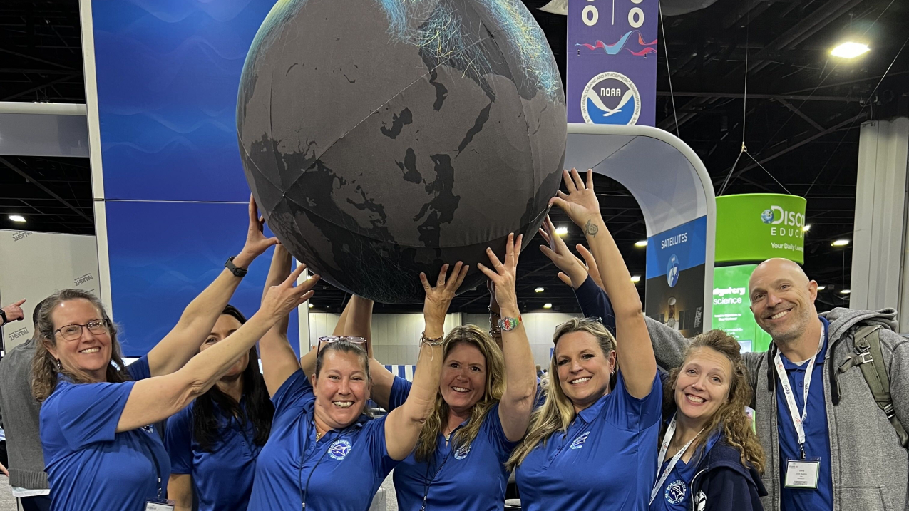 Seven smiling adults in matching blue polo shirts hold up a three-foot wide globe above their heads. The globe has light gray land and dark gray water and colorful lines that indicate hurricane tracks across the globe. They are standing in front of an exhibit booth with the NOAA logo behind them. 