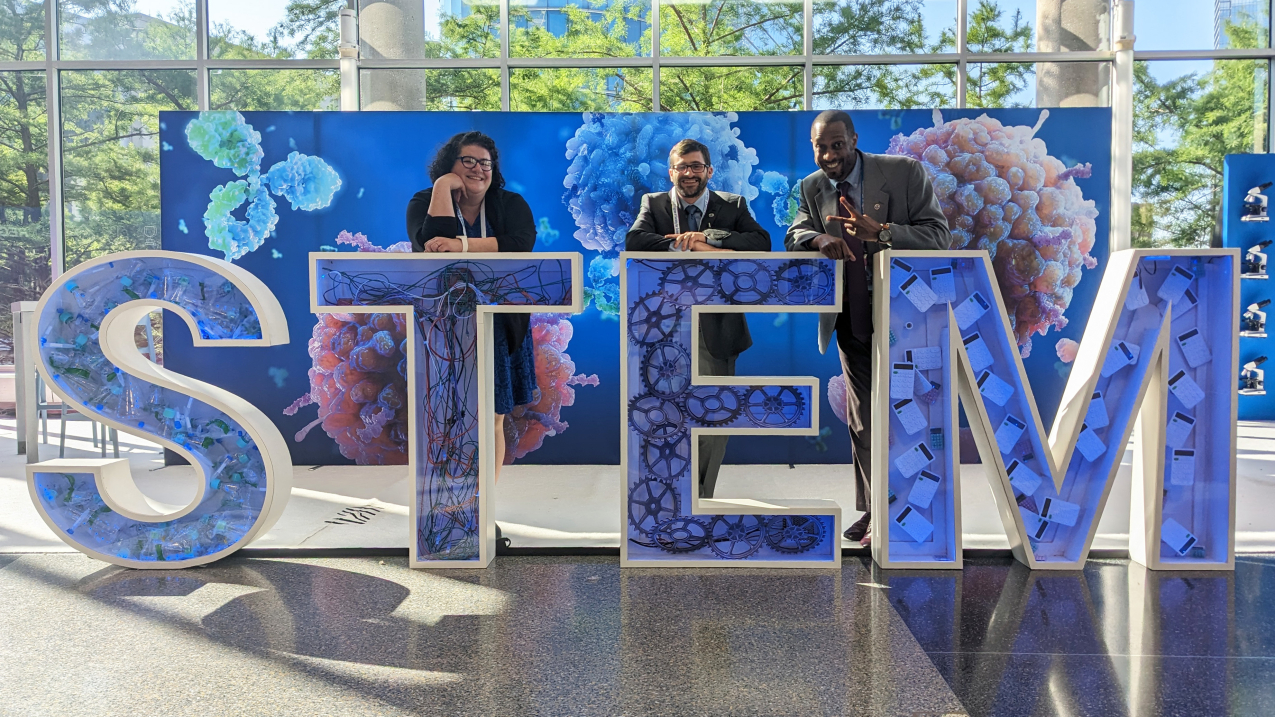 Three smiling adults pose behind 4-foot tall letters that spell out STEM. Each letter has scientific equipment inside of it and there are large printouts of microbes in the background of the picture.