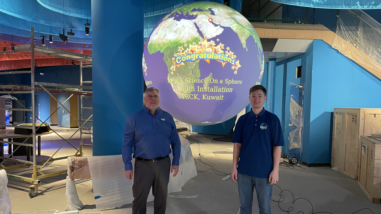 Two men stand in front of a Science On a Sphere globe that displays a world map and the word ‘congratulations’. The globe hangs in an unfinished museum gallery, with parts and boxes on the floor around the site.