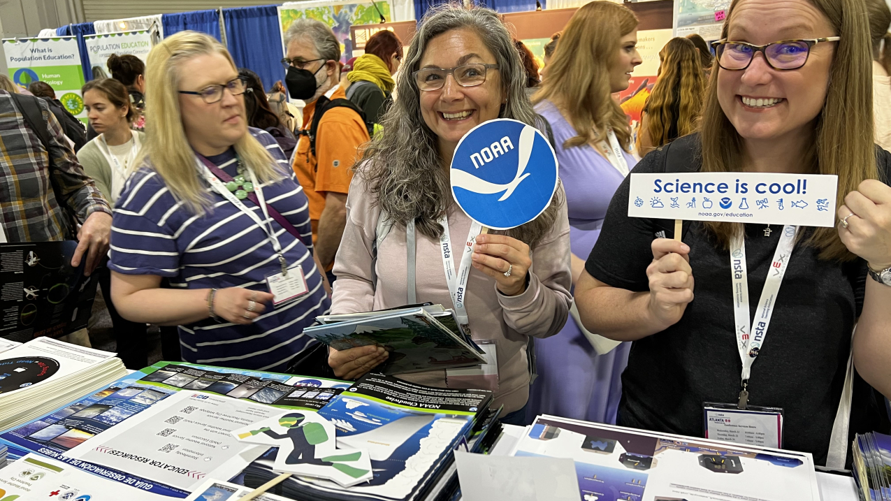 Two smiling teachers hold up signs, one with the NOAA logo, the other reads "Science is cool!," while standing in front of a table covered with educational materials. Other teachers are milling about and visiting other booths behind them.