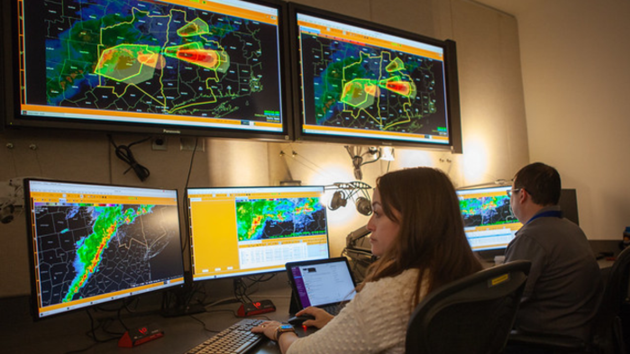 Two people sit at a desk and concentrate on at least six monitors that are displaying different types of weather patterns and data.