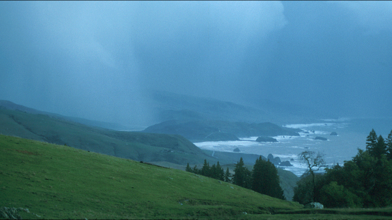 A photograph of a hilly beachfront landscape with heavy rainfall in the background.
