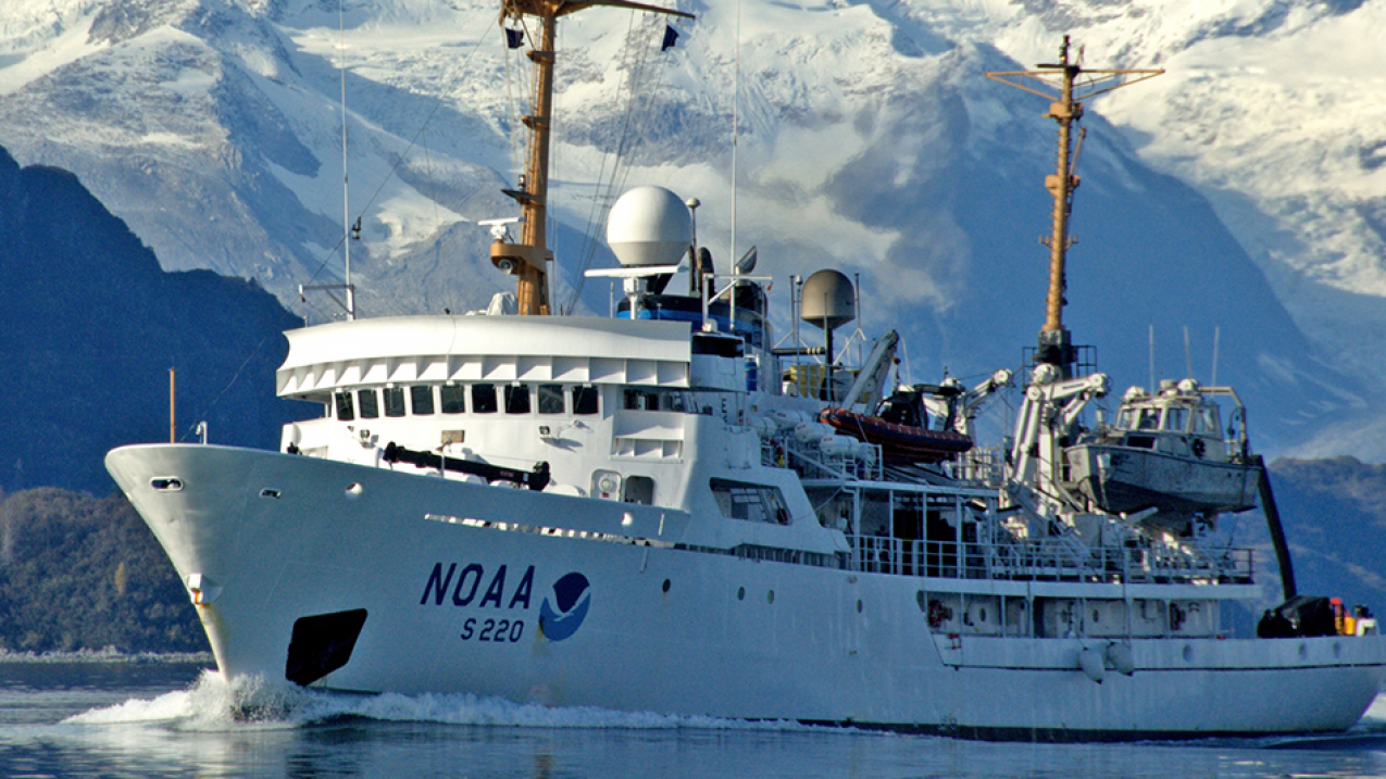A 231-foot hydrographic survey vessel with the NOAA emblem on its bow plows through the water with snow-covered mountains in the background.