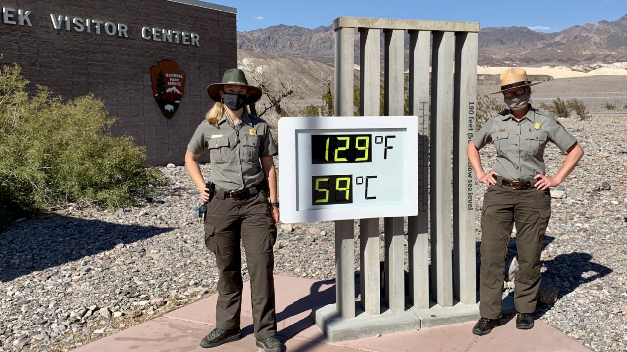 Two rangers in uniform wearing surgical masks stand on either side of a large digital thermometer sign outside the front of Furnace Creek Visitor Center at Death Valley National Park. The thermometer is shown with a high heat reading of 129°F at 4 pm PT on July 12, 2020.