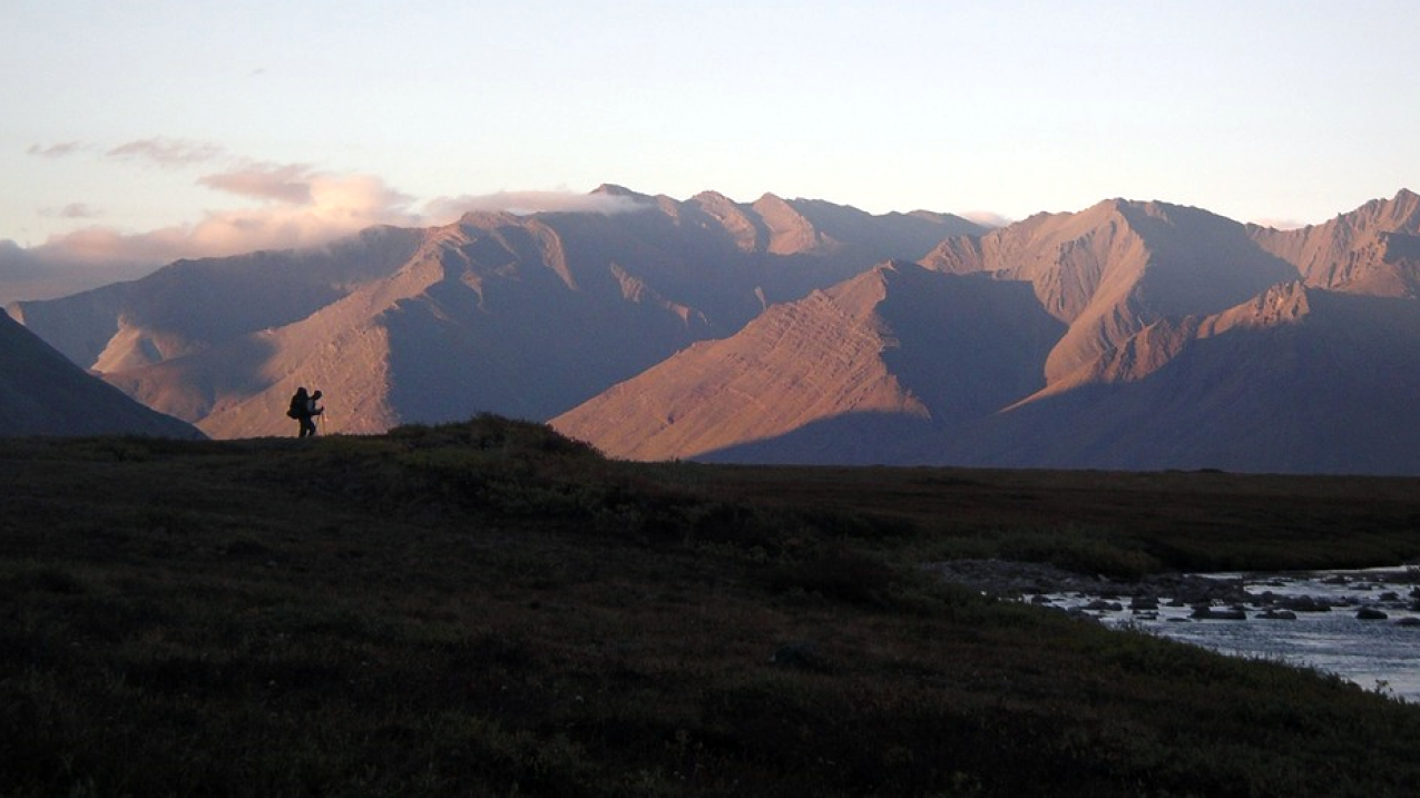 A lone hiker in the distance walks toward a creek with a snowless mountain range in the background. Location: Gates of the Arctic National Park and Preserve. Posted on the park's Facebook page on June 30, 2020.(facebook.com/GatesOfTheArcticNPS)