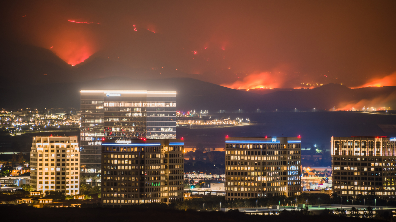 Irvine, California: Two fires began early morning of October 26, 2020, which quickly spread over 30,000 acres in 48 hours. More than 90,000 people were mandated to evacuate. The wildfires in this photo are captured raging in the hills just behind the nighttime urban city landscape of two tall towers and many buildings. The U.S. West experienced its most active wildfire season in 2020, with California recording 5 of the 6 largest wildfires in its history.