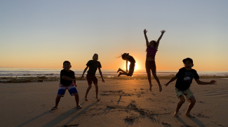 The long shadows of three young boys and two tween girls jumping in the sand with a bright orange sunset behind them making them silhouettes.