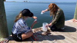Two students sit on edge of dock with water in the background and conduct water quality tesing with bottles and reagents. 