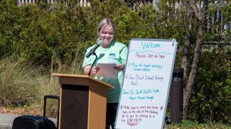 A person stands behind a podium with a microphone outside while holding and looking at papers in her hand. A white board is set up next to her that reads “Welcome please join us here” and lists a schedule of the event. In the background, there are trees and bushes and a few people standing on a wooden deck. 