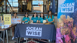 Five people sit behind a table that is draped with a navy blue table cloth that reads “Ocean Wise.” Various papers and animal bones are displayed on the table. To the left is another table with multiple signs. To the right of the table is a long banner with an underwater photo of colorful coral that reads “Ocean Wise.” In the background, there is railing covered by glass and clothes on display for sale. 
