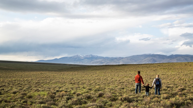 A man, woman, and child walk in an open field in Idaho with dry brush and mountains in the background. April 2021.