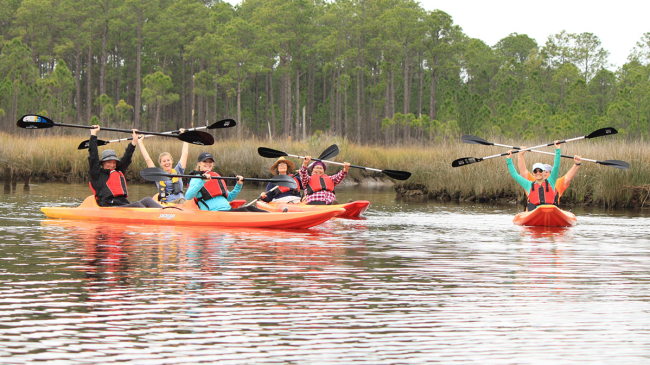 Smiling people in orange kayaks in estuary with raised paddles