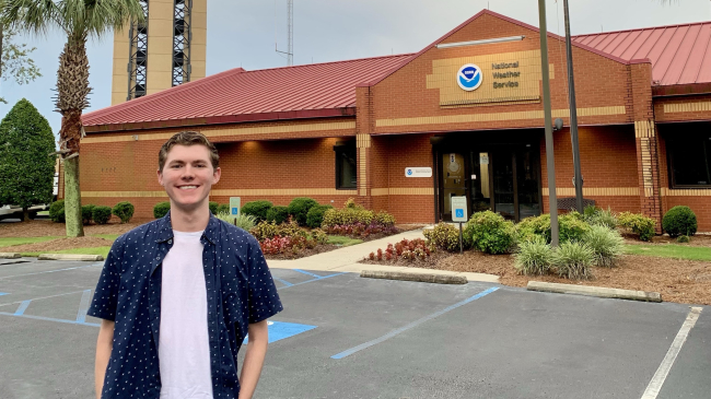 Connor poses in front of a building that has a NOAA logo and the words "National Weather Service" above the door. 
