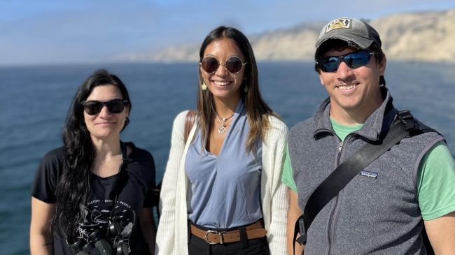 Tammy, Rose, and Jefferson stand together and pose for the camera with relaxed smiles. They wear each sun glasses and stand in front of a water and sky that are calm and blue.