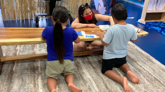 Two children facing away from the camera lean in front of a bench and work on small jigsaw puzzles. From the other side of the bench, Abigail helps one of the children, gesturing to a piece in front of them. She wears a fabric mask over her mouth and nose, as advised by the CDC during the covid-19 pandemic.