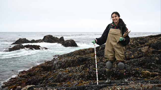 Sofia stands on a rocky shore characteristic of the Pacific coast that is covered in seaweed and mussels. Choppy water breaks against it. She smiles at the camera, wearing a jacket, waders, and knee pads and holds a clipboard in one gloved hand and rests the other on a sampling pole.