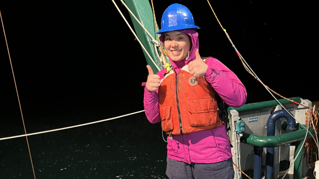 Anna smiles and gives a double thumbs while standing on a vessel at sea. She wears a safety helmet and life jacket and looks energetic and alert despite the dark night sky.