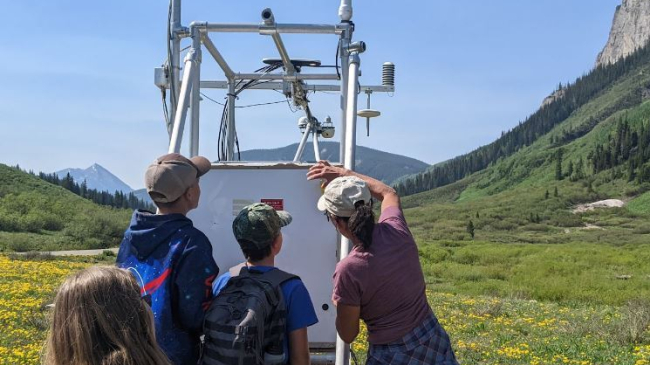 Three children and an adult examine a large metal structure mounted with electronic devices. They are standing in a wide alpine valley filled with wildflowers and surrounded by high mountains.