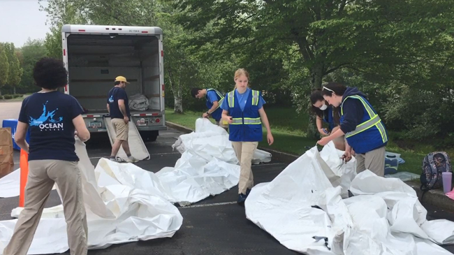 Six people are working outside in a parking lot next to a line of trees. Three people are holding onto large pieces of shrink wrap that are laid out on the parking lot. There is a truck parked behind the group of people with a ramp for loading.
