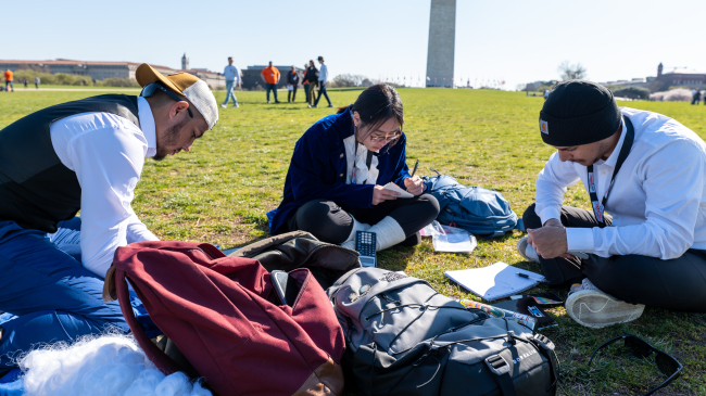 Eric Lopez, Kathlyn Nguyen, and Omar Madrigal make calculations for one of the practical tests in the National Society of Professional Surveyor Student Competition. They sit on the ground with the Washington Monument behind them. Nguyen wears a blue velvet period coat and powdered wigs lie on the ground next to them.