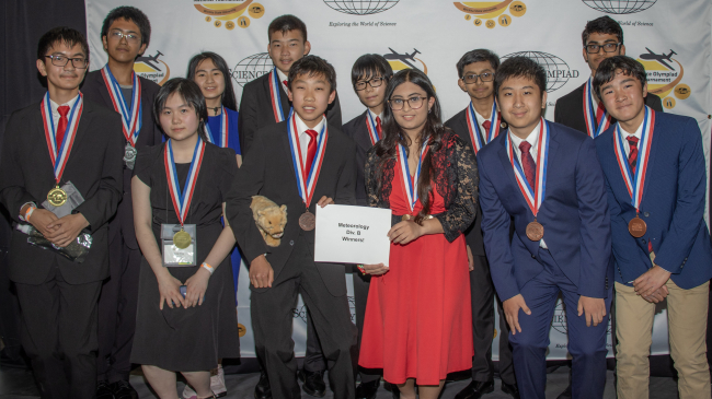 Twelve students dressed in formal attire pose for a group photo in front of a backdrop that has the Science Olympiad National Tournament logo on it. The students in the front row are holding a sign that says Meteorology Div B Winners!