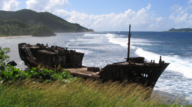 A wrecked wooden ship sits upright on a shoreline.
