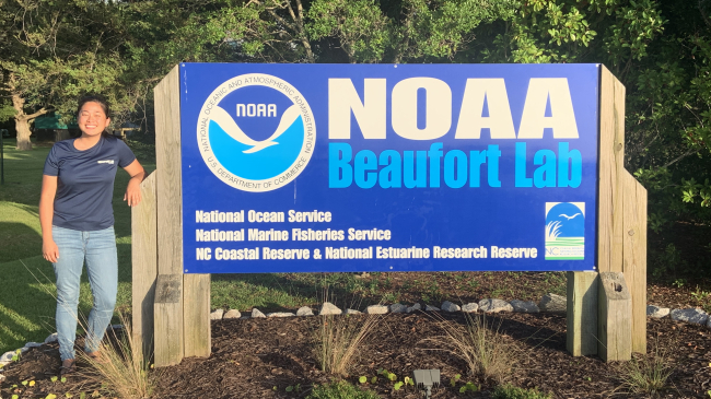 Michaela stands next to a large sign that says "NOAA Beaufort Lab" beside a NOAA logo. Smaller text reads "National Ocean Service, National Marine Fisheries Service, NC Coastal Reserve and National Estuarine Research Reserve