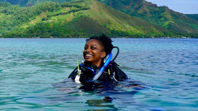 Janelle grins in full SCUBA gear, floating at the surface of crystal clear, blue ocean water. In the background, mountains covered in green vegetation meet the shore.