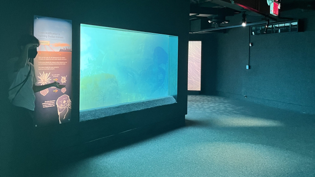 Isaac stands next to an ocean acidification display in an empty room. He holds his hand over his eyes and shrugs the other hand as if looking around in confusion.