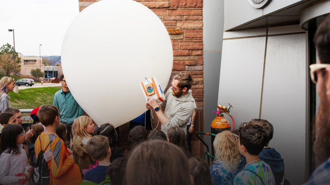 Patrick Cullis, NOAA scientist, surrounded by a sea of enthusiastic children outside the NOAA Boulder building. Boulder's scenic backdrop enhances the enchantment as they eagerly observe the balloon launch, fostering a love for science in young hearts.