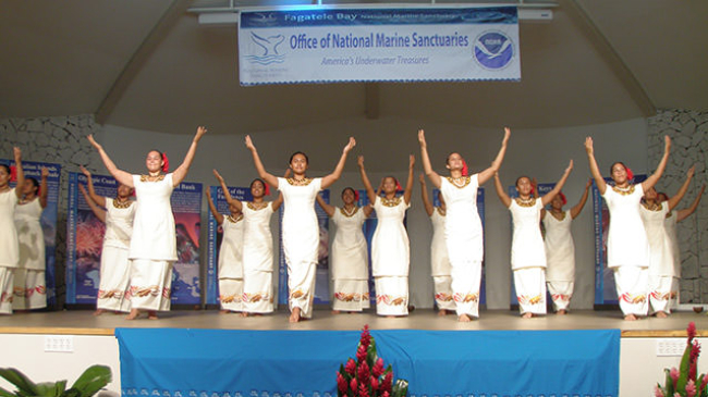 Samoan dancers lift their arms on stage below a National Marine Sanctuary sign.