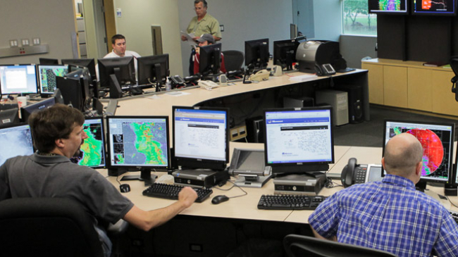 Several meteorologists sitting in front of many computer monitors while forecasting the weather.