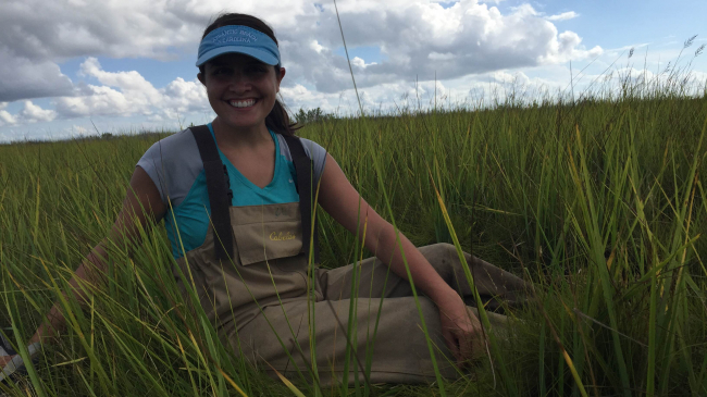 Kim Hernandez wearing waders and sitting among the reeds in a marsh.