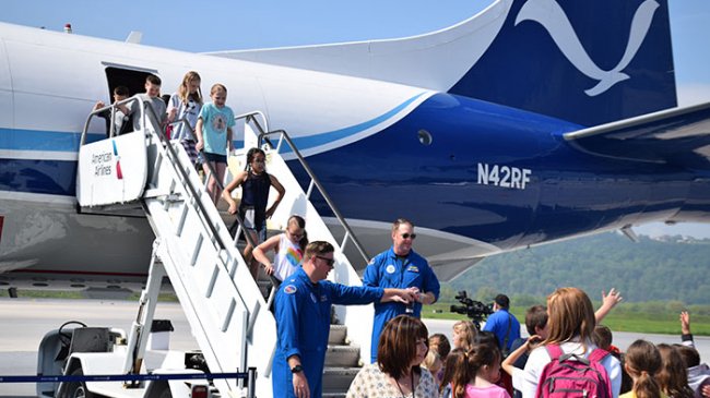 Elementary students climbing down stairs from a NOAA plane. Two NOAA crew are on the ground with other students and teachers.