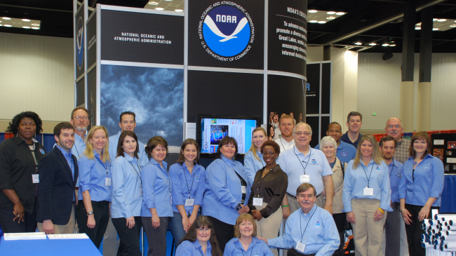 A group of around 20 people stand in front of a large exhibit booth displaying the NOAA logo. 