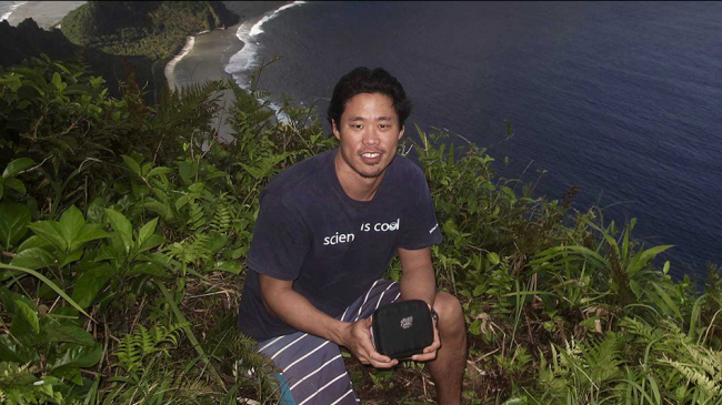 A NOAA employee crouching atop a mountainous island in front of an ocean while holding a small box.