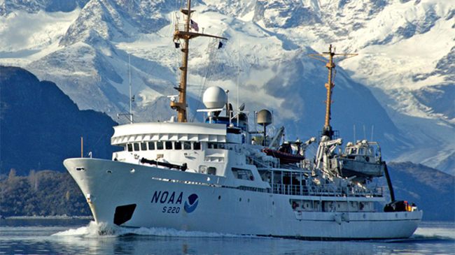 A NOAA hydrographic survey vessel plows through the water with snow-covered mountains in the background.